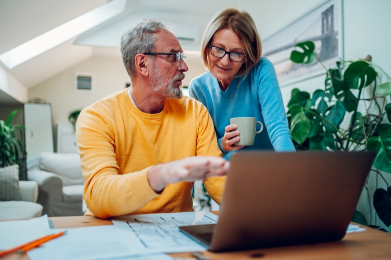 A man sitting at a table with a computer and papers, talking to a woman standing next to him about the uncertain markets. 